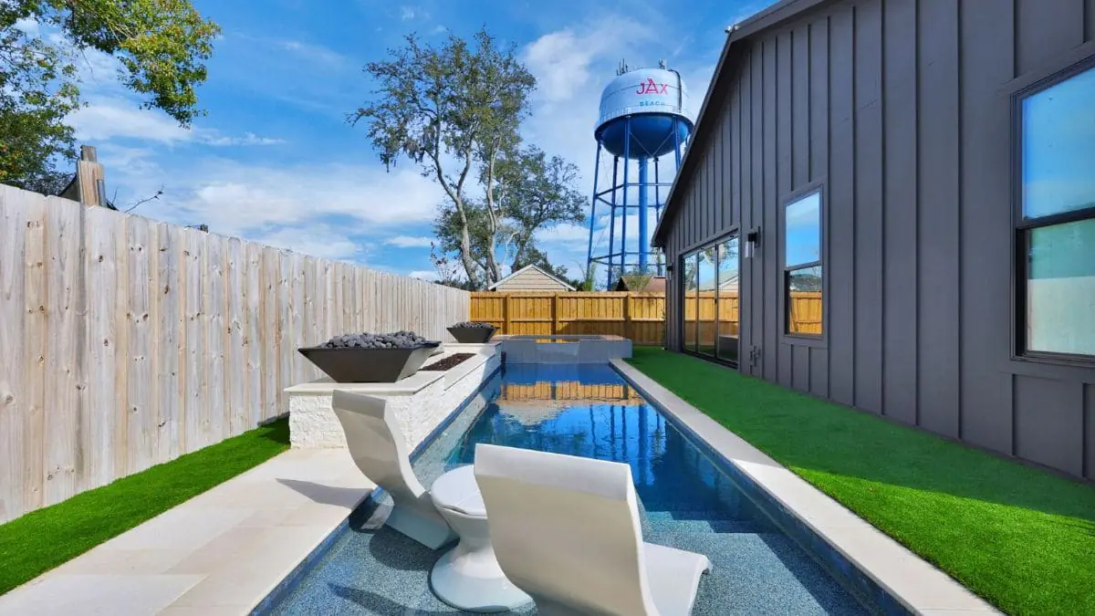 Narrow backyard pool with in-pool lounge chairs, stone wall with fire bowls, artificial turf, and a water tower in the background