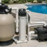 Pool equipment pad with a tan sand filter, black pool pump, white PVC plumbing, and an inground pool with a waterfall in the background