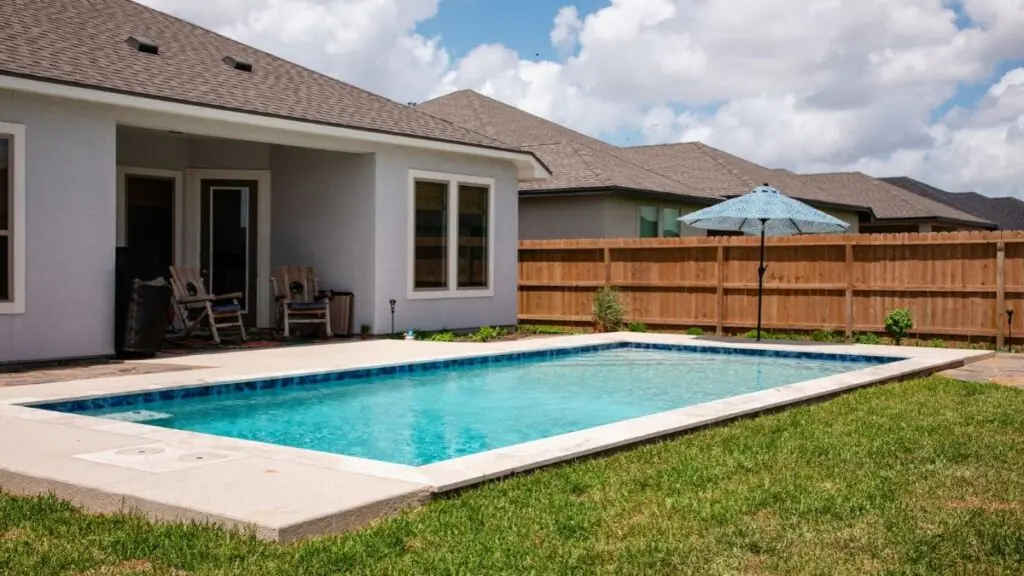 Rectangular backyard pool with a blue tile border, light concrete deck, a blue patio umbrella, and a covered back patio with rocking chairs