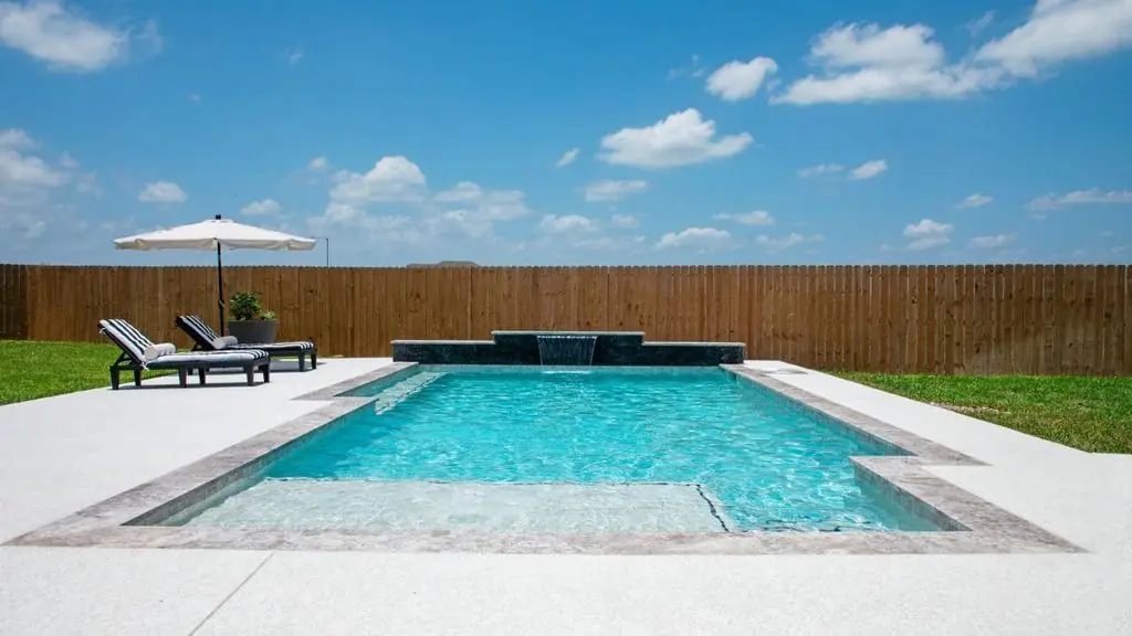 Rectangular backyard pool with a dark stone spillway waterfall, a tanning ledge, light concrete deck, striped lounge chairs, and a patio umbrella beside a wood privacy fence