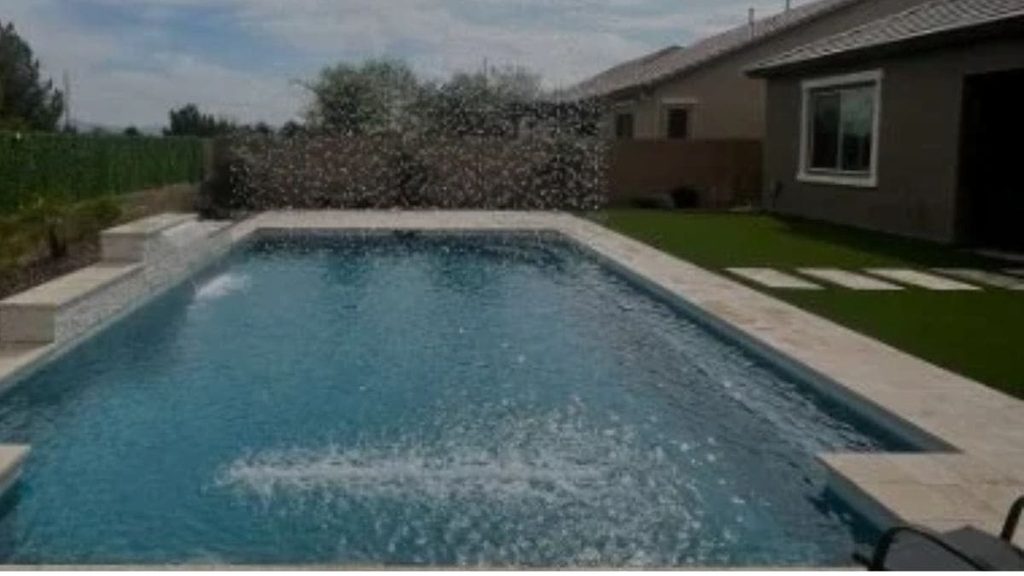 Rectangular backyard pool with water jets and a raised stone water feature, travertine coping, and artificial turf beside a stucco home
