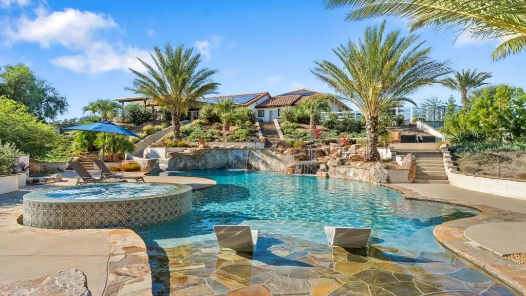 Resort-style backyard pool with rock waterfalls, a round tile spa, a flagstone tanning ledge with in-pool lounge chairs, palm trees, and a hillside home in the background