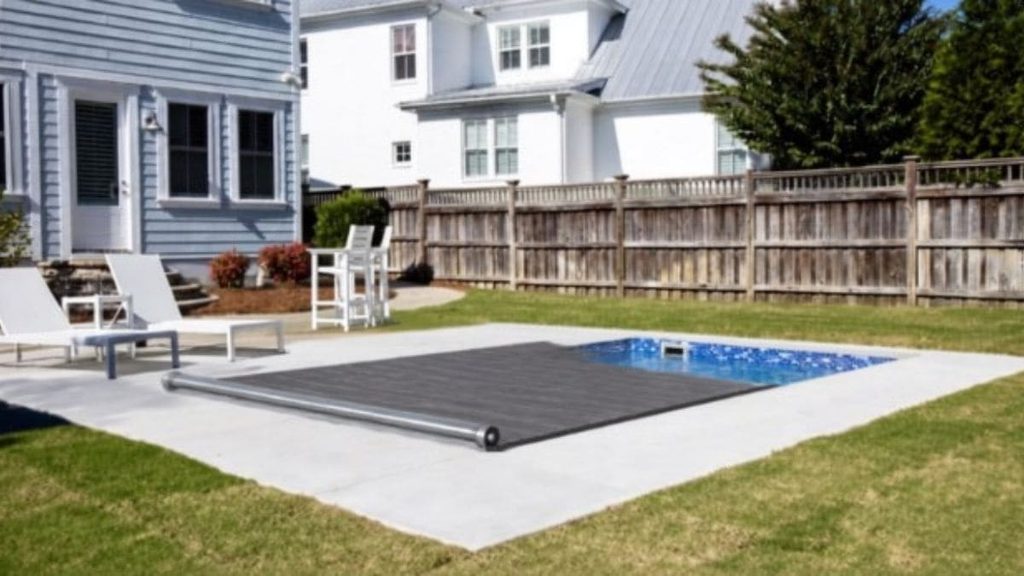 Small backyard pool with a dark automatic safety cover partially retracted on a roller, light concrete deck, white lounge chairs, and a wood privacy fence