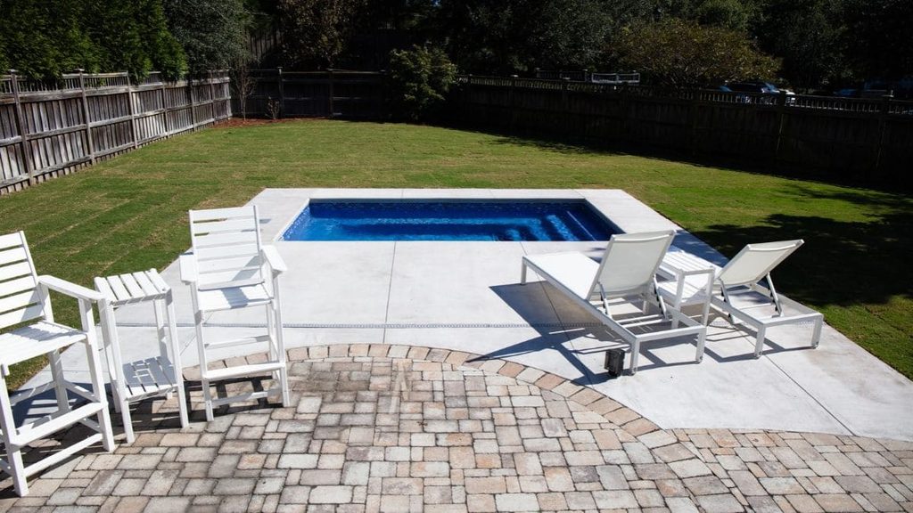 Small backyard pool with a light concrete deck, two white lounge chairs, white high-top chairs, and a curved brick paver patio beside a grassy yard