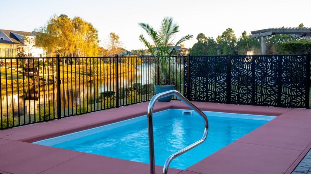 Small rectangular backyard pool with a stainless steel handrail, red-tinted concrete deck, black metal fence, and a pond view at sunset
