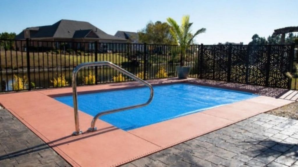 Small rectangular backyard pool covered with a blue solar blanket, stainless steel handrail, red-tinted concrete deck, and a black metal fence with a decorative panel