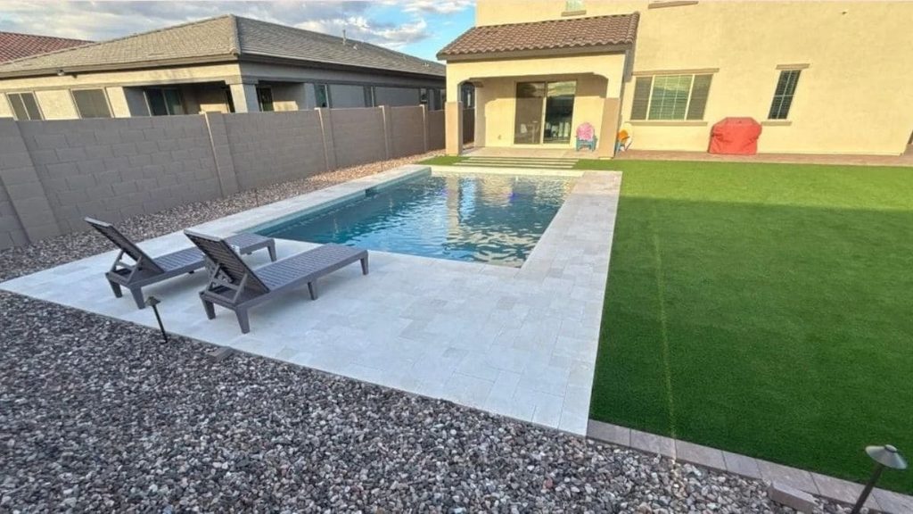 Small rectangular backyard pool with a light travertine deck, two lounge chairs, gravel landscaping, and artificial turf beside a stucco home