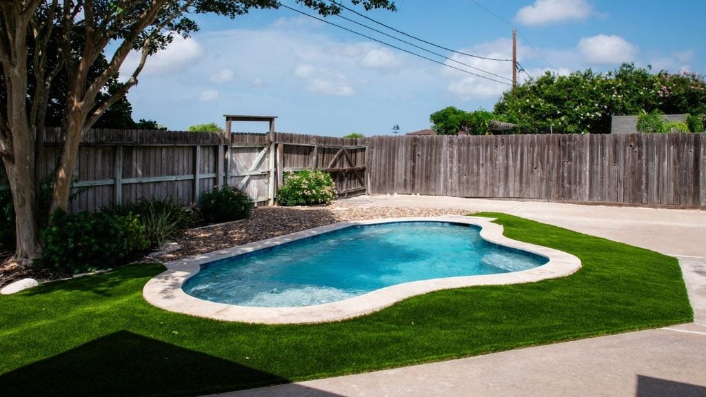 Small freeform backyard pool with travertine coping, artificial turf surround, river rock landscaping, and a wood privacy fence with a gate