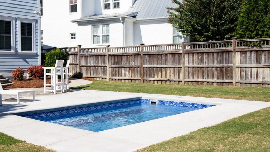 Small rectangular backyard pool with a blue mosaic tile border, concrete deck, white patio chairs, and a wood privacy fence