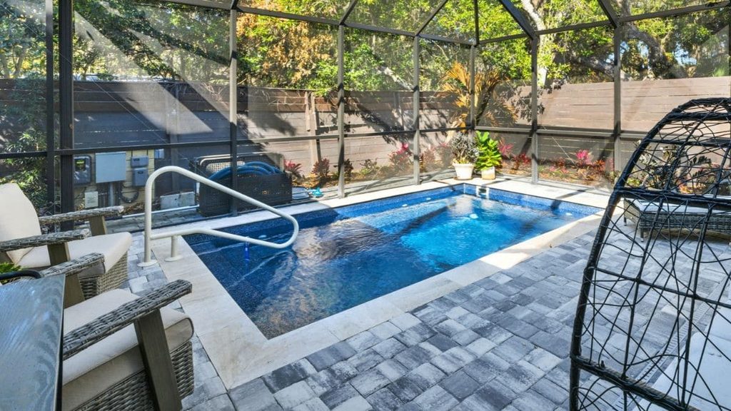 Small inground pool inside a screened enclosure with a handrail, gray paver deck, wicker patio chairs, and tropical plants