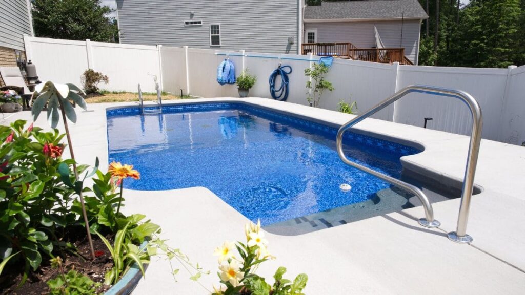 Backyard vinyl liner pool with a blue patterned liner, light concrete deck, stainless steel handrail, flower planters, and a white vinyl privacy fence